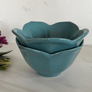 May include: Two teal ceramic bowls stacked. The bowls have a petal design around the rim. A small bouquet of purple and maroon flowers is visible in the background.