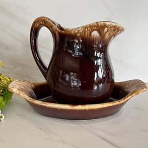 May include: A brown ceramic pitcher with a matching dish. The pitcher has a curved handle and a spout. The dish is oval-shaped with a slightly raised rim. The set has a glossy finish with a two-tone brown and tan color scheme.