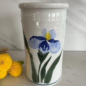 May include: White ceramic vase with a blue and yellow iris flower painted on the side. The vase has a slightly rounded shape and a blue and white stripe around the bottom.