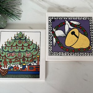 May include: Two ceramic tile coasters with Christmas-themed illustrations. The first coaster features a green Christmas tree decorated with candles, ornaments, and a gingerbread man. The second coaster features a purple background with a white dove holding a gold bell.
