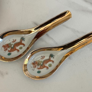 May include: Two white ceramic soup spoons featuring a metallic gold rim and a detailed orange dragon illustration in the center of each bowl. The spoons have a long, curved handle and a deep, rounded basin, showcasing a glossy finish throughout.