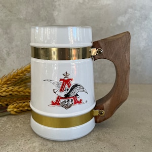 May include: White ceramic beer stein with a gold rim and a wooden handle. The stein features a red eagle logo with the letter "A" and a star.