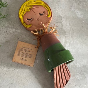 May include: A ceramic wind chime with a brown face and yellow hair. The face has closed eyes and a smile. The chime is attached to a green and brown pot with a straw-like material around the top. The chime has a tag that reads "Unconditional Guarantee - People Care - If for any reason this person refuses to love people, please return it prepaid to our factory. We promise to chastise it severely! When it is properly loving, we will return it to you, perfectly well-adjusted, it will adequately perform its duty of loving people! Thank you - The management - © 1968 by Pacific Stoneware, Inc. U.S.A."