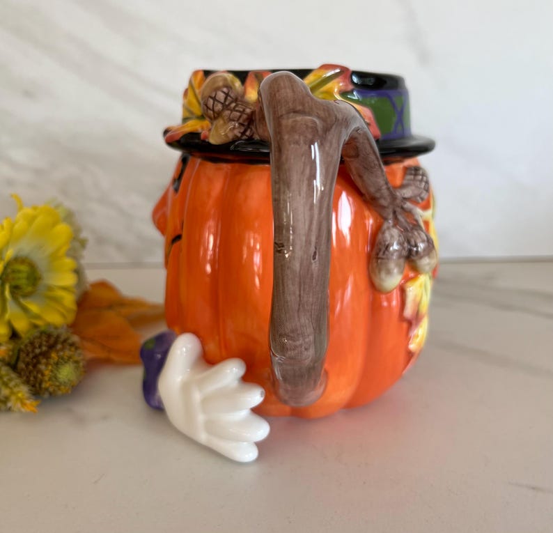 May include: A ceramic pumpkin-shaped mug with a brown handle and a black hat. The mug is orange with a white hand and a purple cuff. The hat is decorated with autumn leaves and a pinecone.