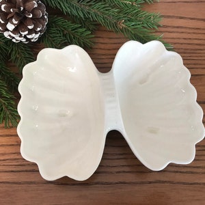 May include: A white, two-section serving dish with a scalloped edge, resembling a bow tie. The dish is made of ceramic or porcelain and sits on a wooden surface, with pine needles and a pine cone in the background.