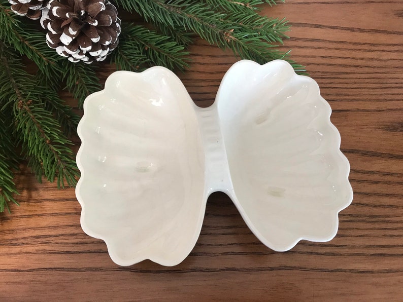 May include: A white, two-section serving dish with a scalloped edge, resembling a bow tie. The dish is made of ceramic or porcelain and sits on a wooden surface, with pine needles and a pine cone in the background.