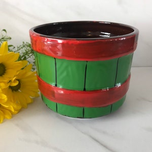 May include: A green and red ceramic planter with a wood grain design. The planter is shaped like a barrel and has a brown interior.