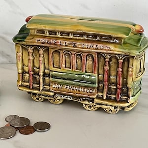 May include: A ceramic cable car bank in shades of green, yellow, and brown, with the words "POWELL & MASON" and "SAN FRANCISCO" visible. The bank has a slot on top for coins and is decorated with red accents. Several coins are scattered in front of the bank.