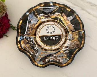 May include: Black glass ashtray with a gold rim and a design featuring various buildings and the text "Expo 67" in a gold circle. The text "Man and His World + Terre des Hommes" is written around the circle.