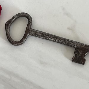 May include: An antique, weathered metal key with a large, oval-shaped handle. The key's shaft and bit show signs of age and wear, with a dark, mottled finish. The key is lying on a white surface.