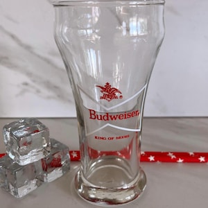 May include: A clear Budweiser beer glass with the brand's logo and text "KING OF BEERS" in red. The glass is empty, with a rounded base and a flared top. Beside the glass are three ice cubes and a red straw with white stars.