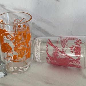 Peut inclure: Deux verres à boire vintage transparents avec des illustrations orange et rouges. Un verre présente une image détaillée d'un homme avec un chapeau et le texte "Davy Fought So Texas". L'autre verre a une illustration rouge d'un homme avec un chapeau et un fusil.