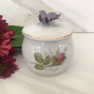 May include: A white porcelain jar with a floral design and a purple flower on the lid. The jar has a gold trim around the top and bottom.