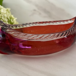May include: A red glass canoe-shaped dish with a clear glass rim and a white rope design. The dish has a iridescent finish.
