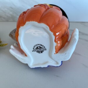 May include: A ceramic pumpkin-shaped creamer with an orange exterior and a black interior. The creamer has a white handle and a blue base. The bottom of the creamer has the text "FITZ AND FLOYD ESSENTIALS".