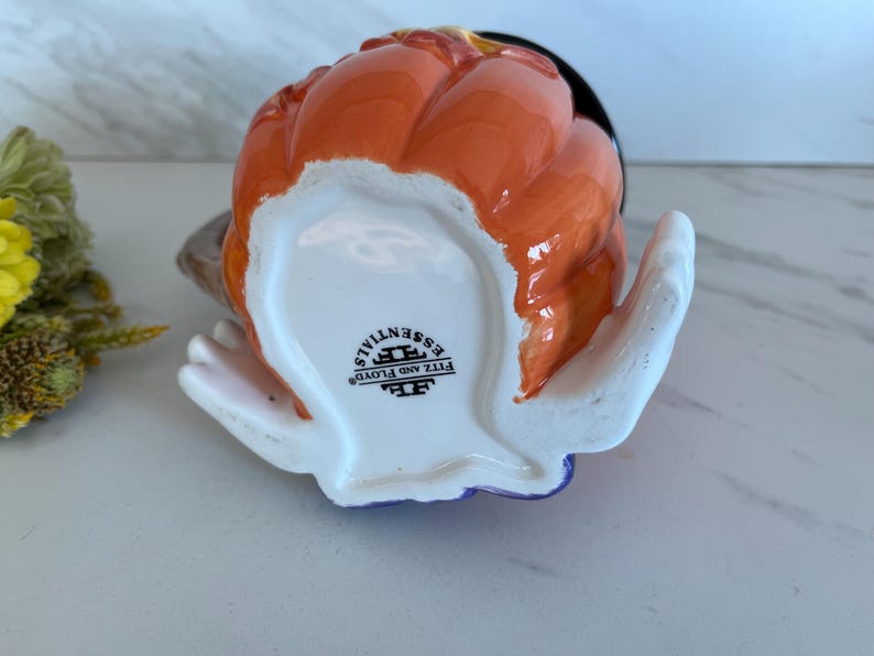 May include: A ceramic pumpkin-shaped creamer with an orange exterior and a black interior. The creamer has a white handle and a blue base. The bottom of the creamer has the text "FITZ AND FLOYD ESSENTIALS".