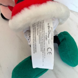 Puede incluir: Un Mickey Mouse de peluche vestido de Pap&aacute; Noel con un traje rojo, ribetes blancos y zapatos verdes. La etiqueta dice "The Walt Disney Company" y "Disney".