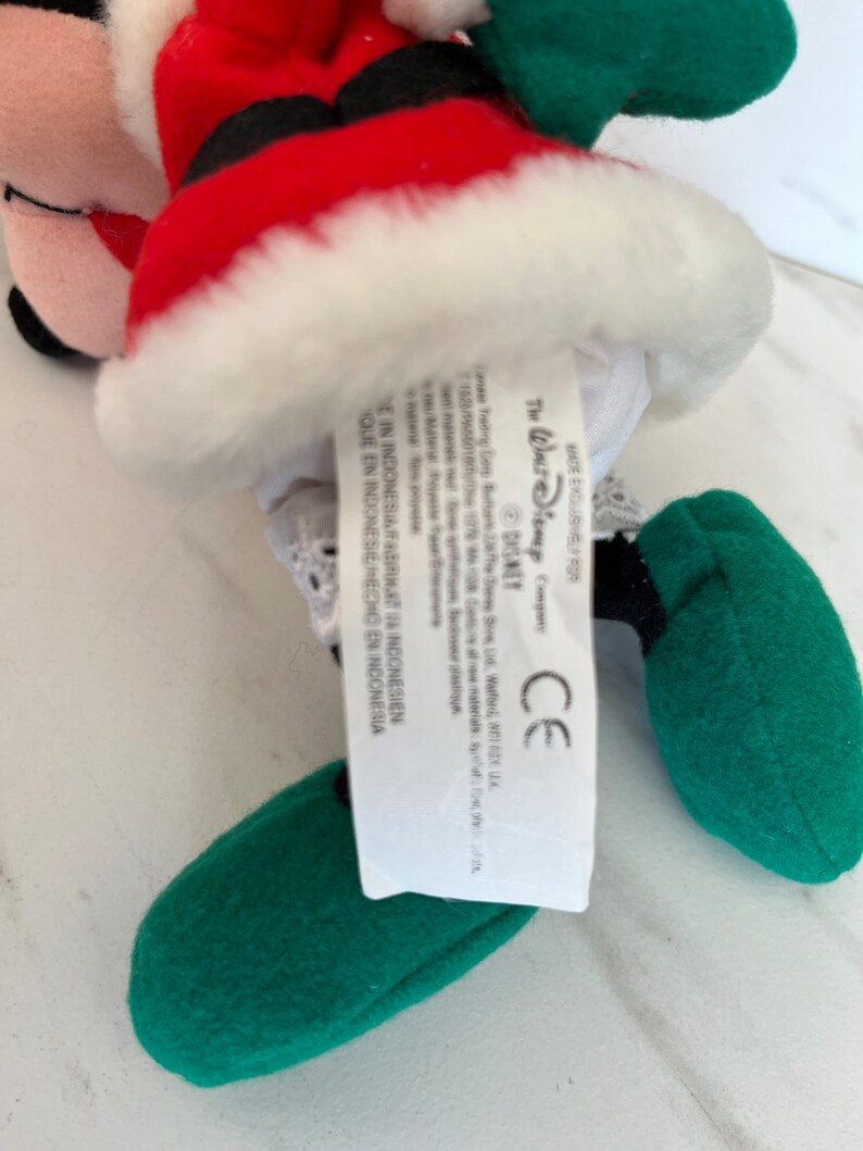 Puede incluir: Un Mickey Mouse de peluche vestido de Pap&aacute; Noel con un traje rojo, ribetes blancos y zapatos verdes. La etiqueta dice "The Walt Disney Company" y "Disney".