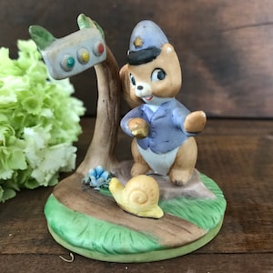 May include: A ceramic figurine of a squirrel wearing a blue police uniform and a hat, holding a traffic light. The squirrel is standing on a green base with a snail beside it.