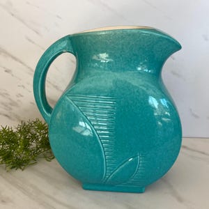 May include: A turquoise ceramic pitcher with a leaf design. The pitcher has a wide mouth and a handle.