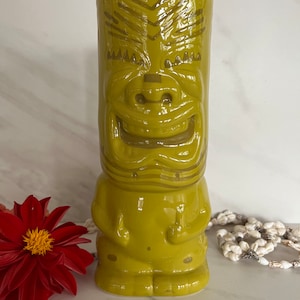 May include: A green ceramic tiki mug with a smiling face and a patterned design. The mug is glazed and has a glossy finish.