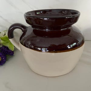 May include: A two-toned ceramic crock with a wide, flared rim and a single side handle. The upper portion of the vessel is finished in a glossy dark brown glaze, while the lower body features a smooth, cream-colored base. The piece is rounded.