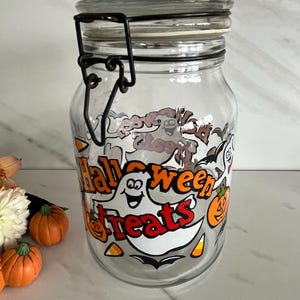 May include: Clear glass jar with a black metal clasp and lid. The jar is decorated with Halloween-themed graphics, including ghosts, pumpkins, and the words "Halloween Treats". The jar is empty.