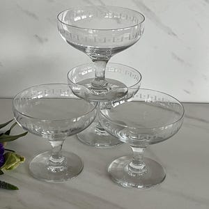 May include: Four clear champagne coupe glasses stacked, with a decorative etched border around the rim. The glasses are on a white surface, with a few purple flowers in the lower left corner.