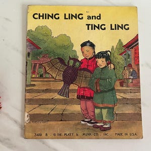 May include: A vintage children's book cover featuring two young girls in traditional Chinese clothing, holding a kite shaped like a bat. The book title is "Ching Ling and Ting Ling" and the copyright information is "3400 B. © THE PLATT & MUNK CO., INC. MADE IN USA."