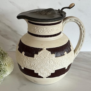 May include: A cream-colored ceramic pitcher with dark brown horizontal stripes and decorative embossed patterns. The pitcher has a handle and a hinged lid with a small wooden knob. The pitcher is a decorative item.