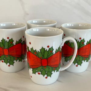 May include: Set of four white ceramic mugs with a Christmas design. Each mug features a green holly wreath with a red bow and red berries.
