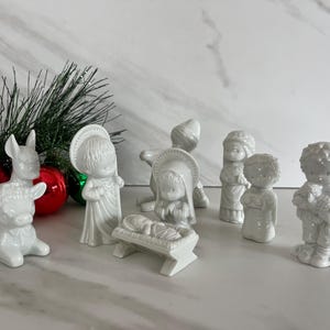 May include: A set of white ceramic nativity figurines including a baby in a manger, figures with halos, a donkey, and a sheep. The collection features smooth, glossy surfaces and detailed facial expressions, arranged together as a traditional holiday display.