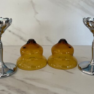 May include: Set of two silver-toned metal candle holders with a decorative, scalloped top and a slender stem. Two amber-colored glass candle covers are also present, creating a vintage aesthetic.