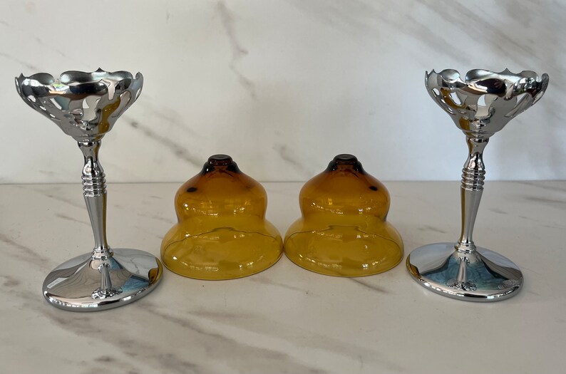 May include: Set of two silver-toned metal candle holders with a decorative, scalloped top and a slender stem. Two amber-colored glass candle covers are also present, creating a vintage aesthetic.