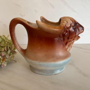 May include: A brown ceramic pitcher shaped like a bull's head with a blue glaze around the bottom. The pitcher has a handle and a spout.