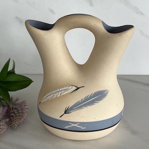 May include: Cream-colored ceramic vase with a unique handle and two openings. The vase features a blue band and two feather designs. The rim and handle opening are painted blue. A decorative piece for the home.