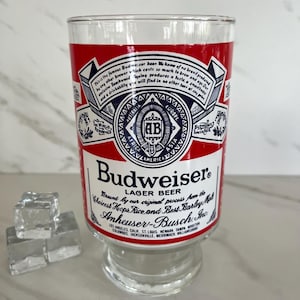May include: A clear Budweiser beer glass with a red and white label. The label features the Budweiser logo, text, and a decorative design. The glass sits on a clear pedestal base. Ice cubes are in the foreground.
