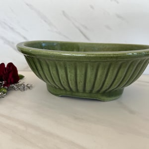 May include: A green ceramic planter with a fluted design. The oval-shaped container has a wide opening and a low, rectangular base. The planter is a muted green color and sits on a white surface. Red flowers are visible on the left.