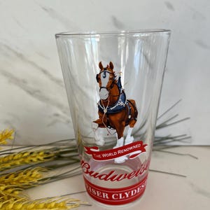 May include: Clear glass pint glass featuring a Clydesdale horse in harness. The glass has a red base with the words "Budweiser" and "The World Renowned" printed on it. The horse is brown and white.