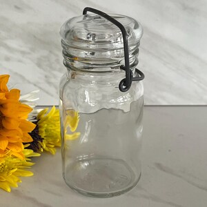 May include: A clear glass jar with a matching glass lid secured by a dark gray metal wire bail closure. The jar has a cylindrical body and a rounded rim, providing a simple and functional design for storage. The glass is transparent and smooth.