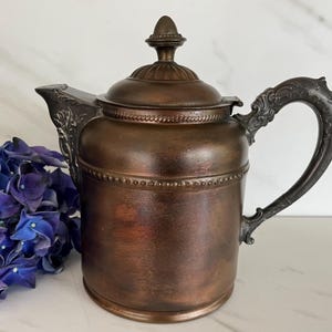 May include: A metallic copper-toned teapot featuring a cylindrical body, a decorative handle with ornate scrollwork, a matching lid with a finial, and a spout adorned with leaf-like detailing. The surface has a dark, weathered patina finish.