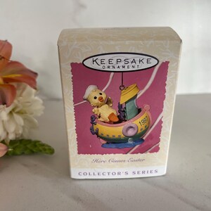 May include: A yellow chick wearing a blue sailor hat sits in a green and yellow boat with a white sail. The boat is on a pink background with the words "Here Comes Easter" and "Collector's Series" printed below. The box is labeled "Keepsake Ornament" and has the year "1997" printed on the boat.