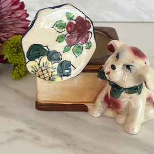 May include: A ceramic trinket box featuring a cream-colored dog figurine with a green neck ribbon. The lid is an octagonal shape decorated with hand-painted red flowers and green leaves. The base of the container is a light tan color with brown trim.