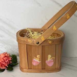 May include: A light brown wooden Easter basket with a handle, decorated with yellow chicks and pink egg designs. The basket is filled with yellow straw. A pink flower with green leaves is in the foreground.
