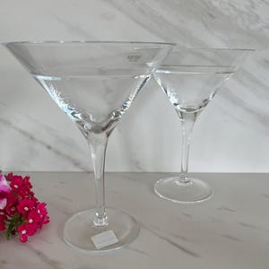 May include: Two clear martini glasses with long stems and wide, triangular bowls. The glasses are made of transparent glass and sit on a white surface. A small bunch of pink flowers is in the lower left corner.
