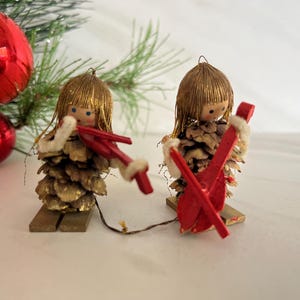 May include: Two vintage Christmas ornaments. The figures are made from pine cones, with wooden heads and red skis. The figures have gold hair and wear white trim. A red Christmas bauble and green pine branches are in the background.