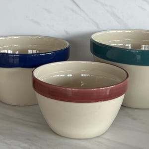 May include: Three ceramic mixing bowls in varying sizes. The bowls are off-white with colored rims: navy blue, teal, and burgundy. The bowls are arranged on a white marble surface. A small bouquet of flowers is visible on the left.