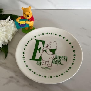 May include: White ceramic plate with a green dotted border and the words "easy breezy days..." in green. The plate features a cartoon image of Winnie the Pooh. A Winnie the Pooh figurine is in the background.