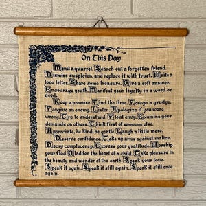May include: A rectangular wall hanging featuring a tan linen fabric with dark blue printed text titled On This Day. The text includes inspirational phrases with decorative initial letters and a floral border. It is held by two wooden rods.