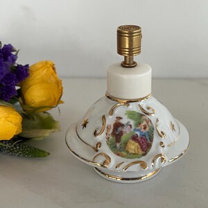 May include: A white porcelain perfume bottle with a gold-colored top and accents. The bottle features a painted scene of a couple in a garden. The bottle has a unique shape and is likely vintage.